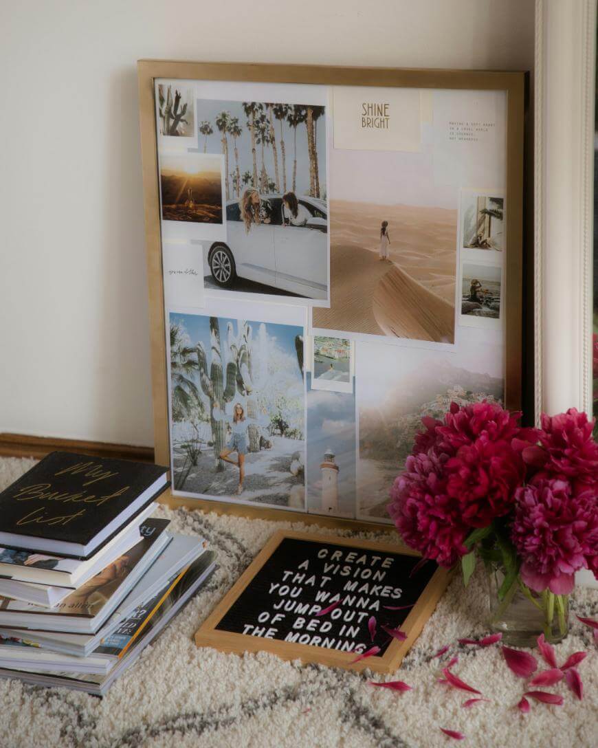 A framed collage of travel photos and inspirational quotes leans against a wall on a carpeted floor. Nearby are stacked books, vibrant pink flowers, and a letterboard displaying a motivational message.