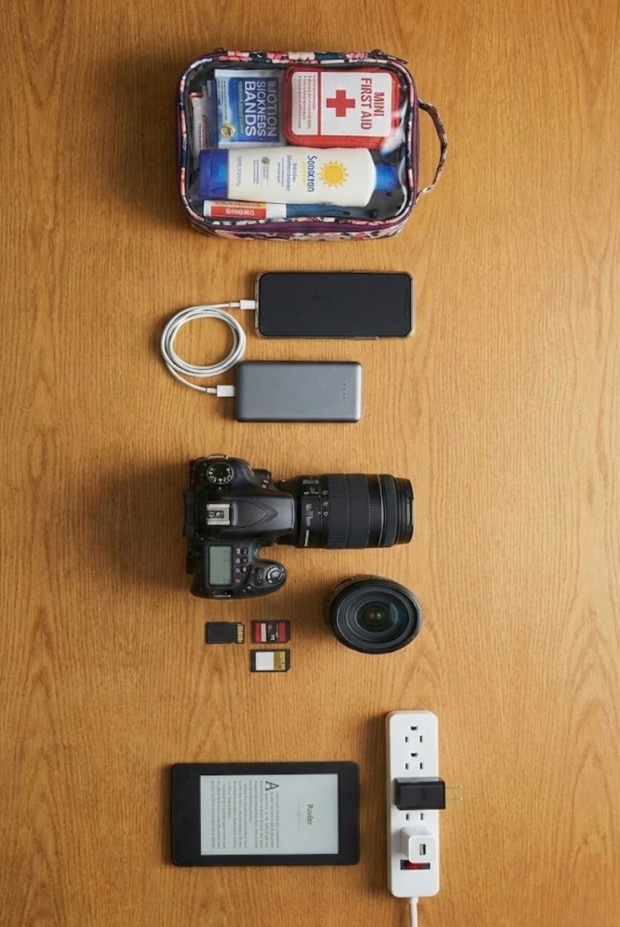 A flat lay of travel essentials on a wooden surface, including a first aid kit, charging cables, power banks, a camera with lens, SD cards, e-reader, extra camera lens, and a power strip.