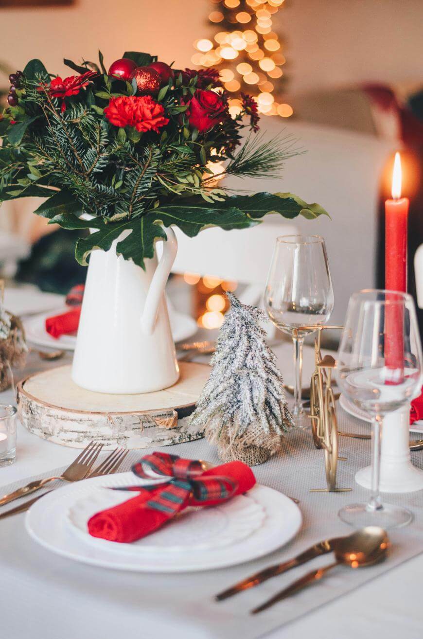 A festive table setting with a white pitcher filled with red flowers and greenery, a red candle, wine glasses, a small snowy tree decoration, and plates with red napkins tied in plaid ribbon. A Christmas tree is blurred in the background.