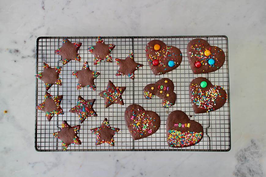 A cooling rack with star, heart, and Mickey Mouse-shaped chocolate cookies decorated with colorful sprinkles and candy pieces, on a light marble surface.