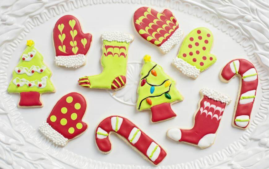 A white plate with decorated Christmas cookies shaped like mittens, stockings, trees, and candy canes. The cookies are iced in red, green, yellow, and white with festive holiday designs.