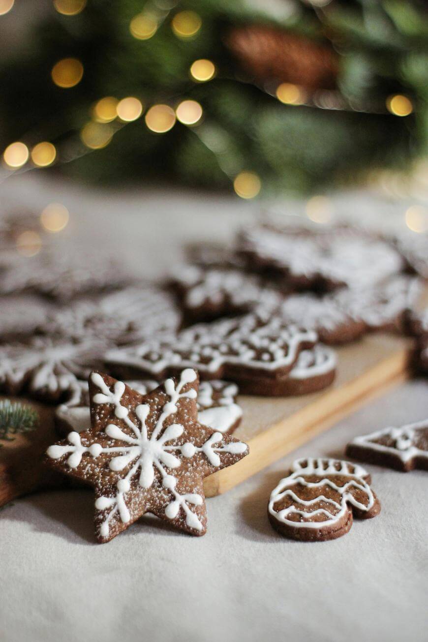 A star-shaped gingerbread cookie decorated with white icing sits in front of more assorted holiday cookies on a wooden board, with blurred festive lights and greenery in the background.