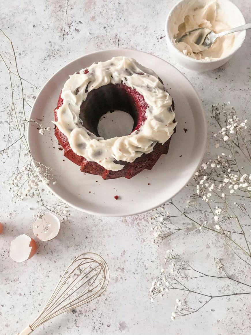 A bundt cake with white frosting sits on a round plate, surrounded by a whisk, eggshells, a bowl of frosting with a spoon, and sprigs of babys breath flowers on a light tabletop.