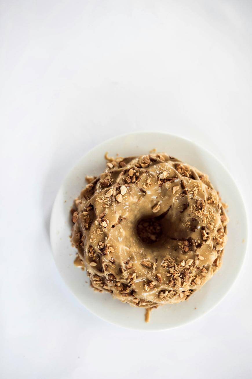A bundt cake topped with caramel glaze and chopped nuts sits on a white plate, viewed from above against a white background.