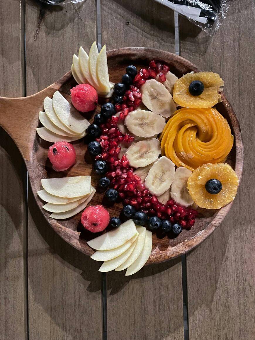 A round wooden platter with sliced apples, watermelon balls, banana slices, pomegranate seeds, blueberries, and decorative orange slices arranged in a floral pattern on a wooden table.