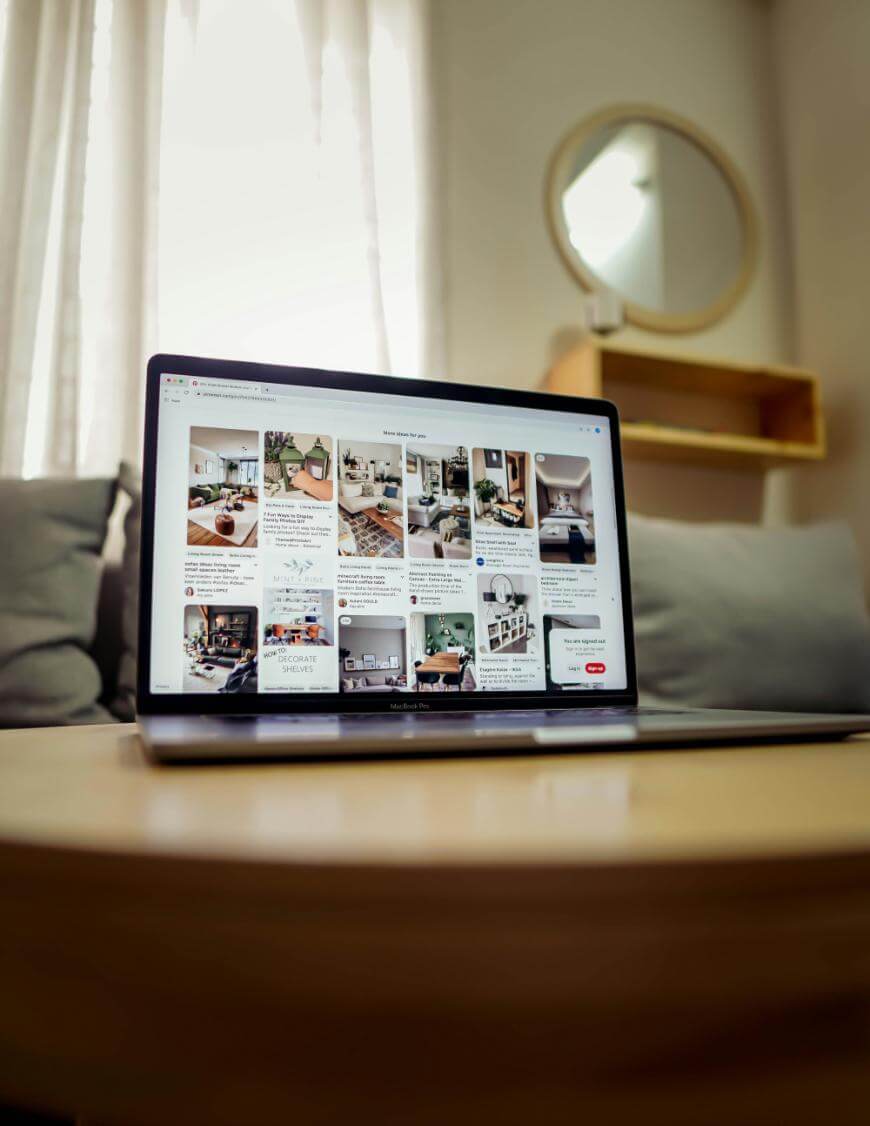 A laptop on a wooden table displays a Pinterest page with home decor ideas. The background features a gray couch, sheer curtains, and a round mirror on the wall.