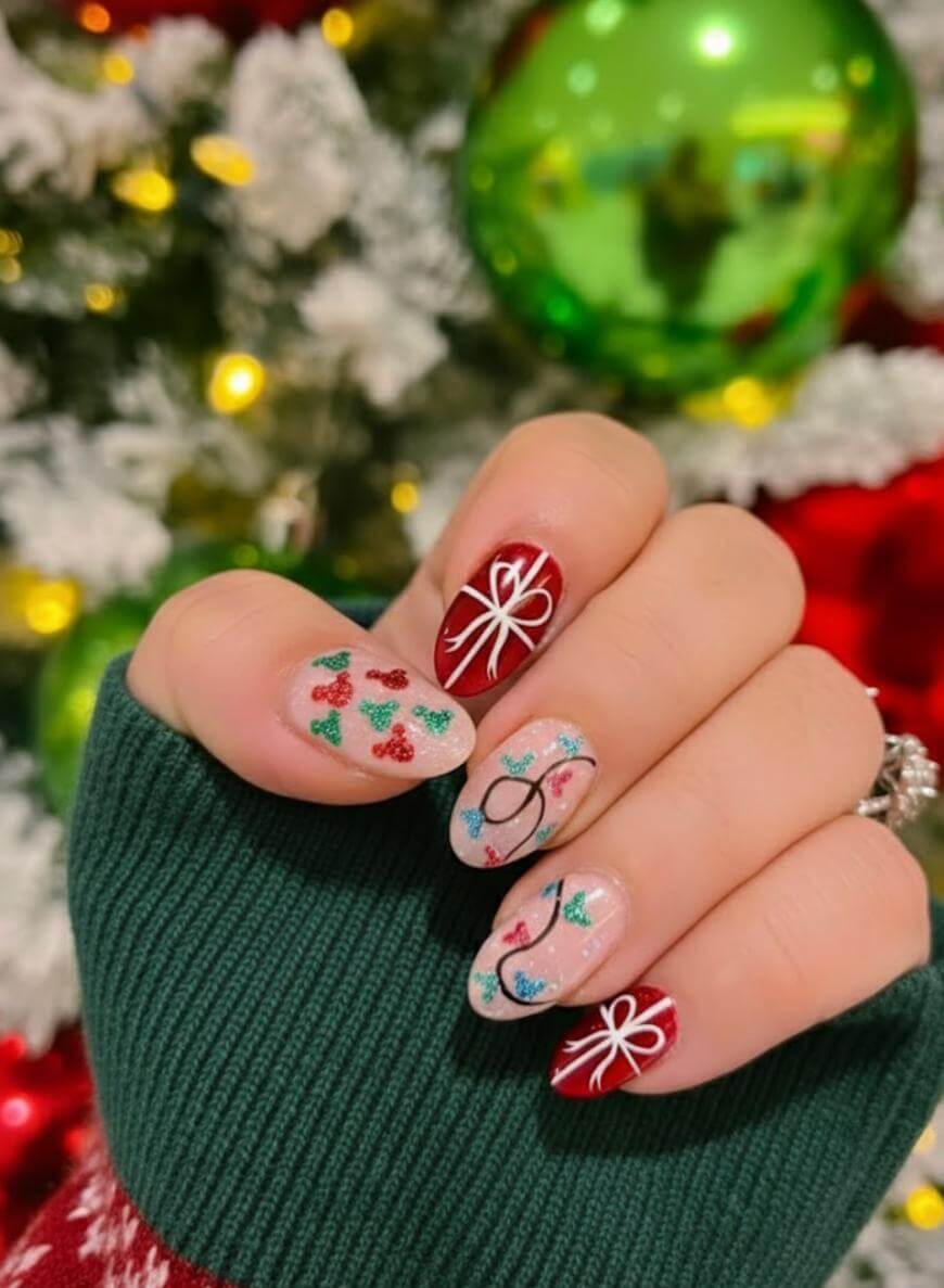 A hand with festive Christmas nail art featuring holly, ornaments, and white ribbon designs is held up in front of a decorated Christmas tree with a large green ornament in the background.