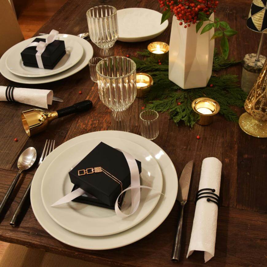 A festive dinner table set with white plates, gift boxes tied with ribbon, glassware, candles, a white vase with red berries, green foliage centerpiece, and neatly rolled napkins with black rings.