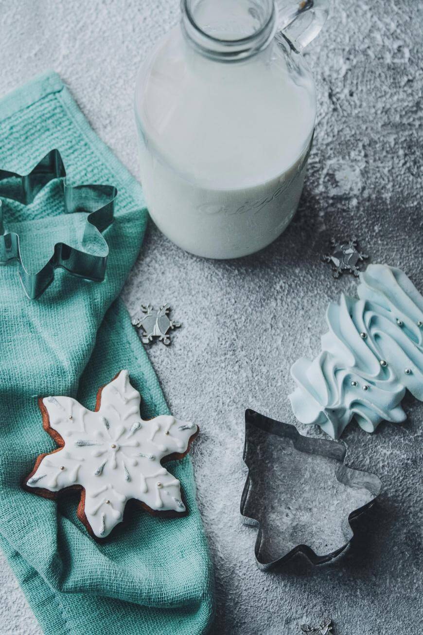 A snowflake-shaped iced cookie, blue napkin, tree-shaped cookie cutter, blue meringues, snowflake decorations, and a glass jug of milk on a textured gray surface.