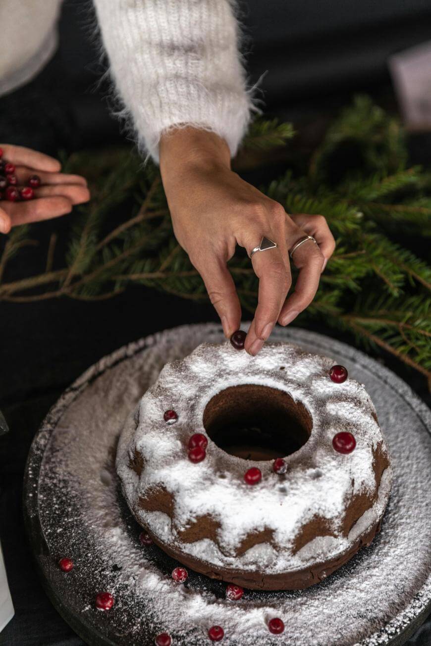 A person decorates a powdered Bundt cake with cranberries, placing them on top. The cake sits on a dark plate, and evergreen branches are visible in the background.