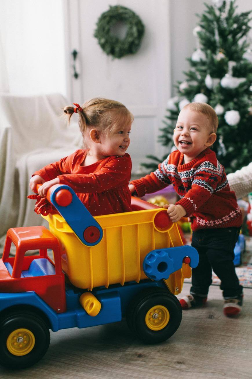 Two young children in festive red outfits play with a large toy dump truck in a living room decorated for Christmas, with a tree and wreath in the background. Both children are smiling and appear happy.