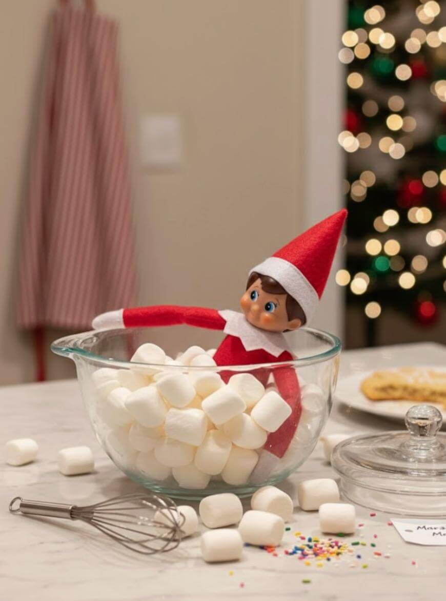 A plush elf doll in a red and white outfit sits inside a glass bowl filled with marshmallows on a kitchen counter, with a whisk and scattered marshmallows beside it. A lit Christmas tree is blurred in the background.