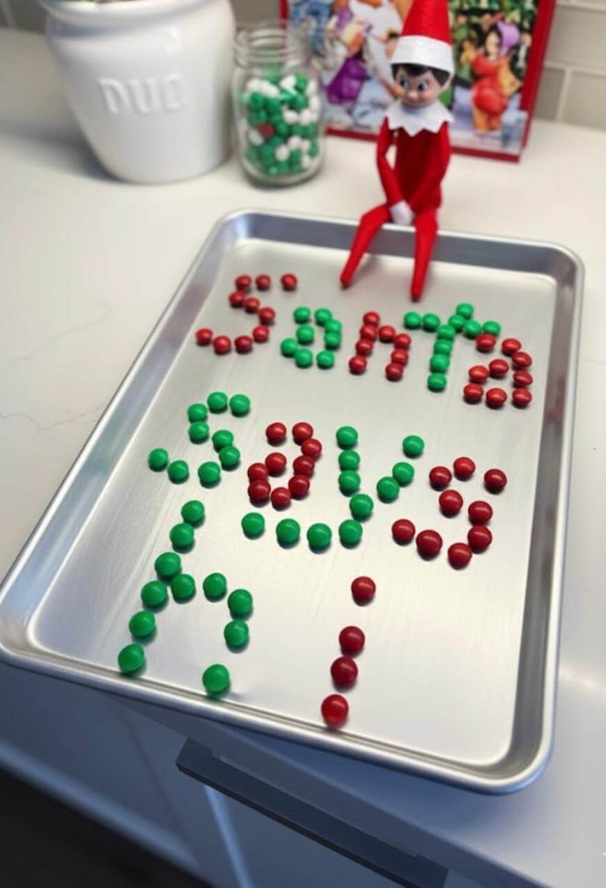 A baking tray on a counter spells “Santa says hi” with green and red candies. An Elf on the Shelf doll sits at the tray’s edge, and jars of candies and holiday decor are in the background.