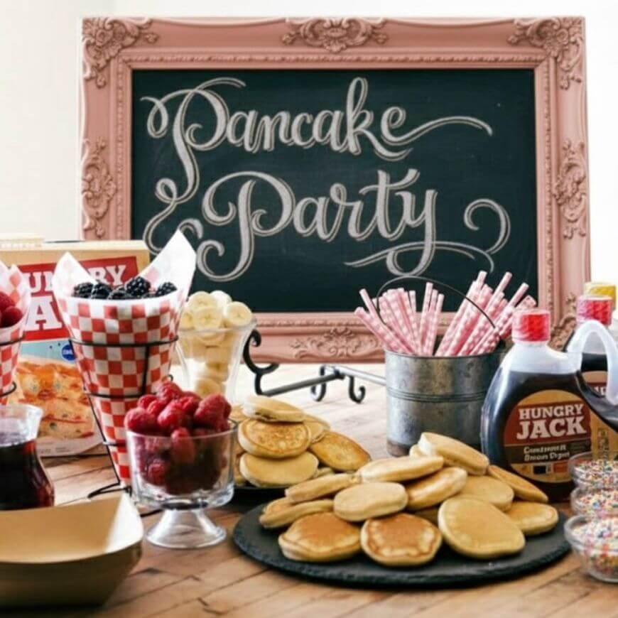 A table set for a Pancake Party with a chalkboard sign, stacks of pancakes, berries, bananas, syrup bottles, checkered cups, and jars of straws. The scene looks festive and inviting.