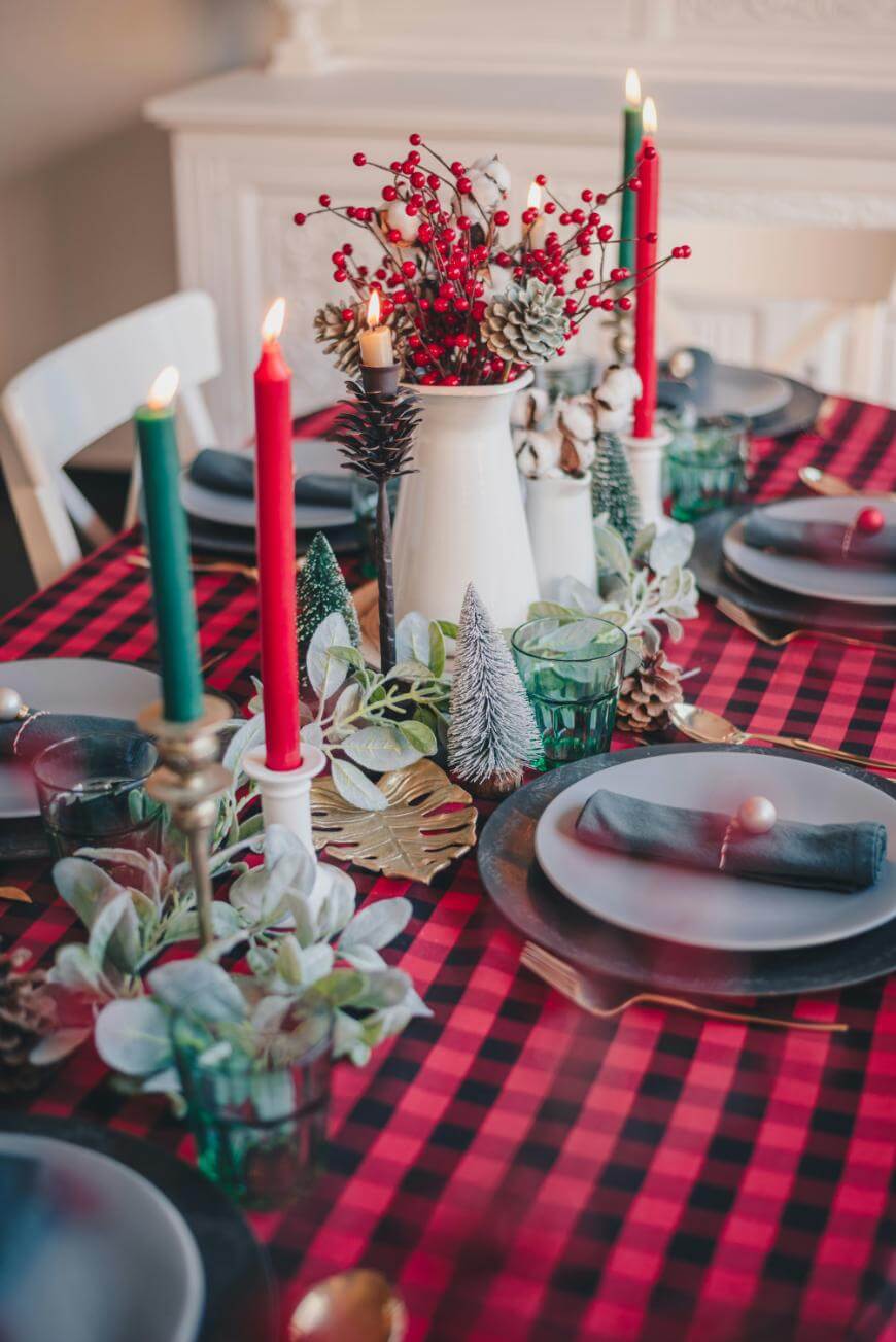 A festive holiday table is set with a red and black plaid tablecloth, green and red candles, small decorative trees, greenery, and a white pitcher with red berries as a centerpiece. Plates and glasses are neatly arranged.