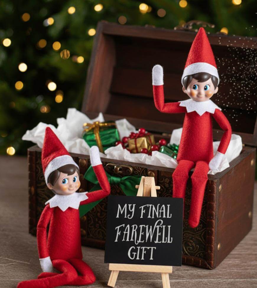 Two Elf on the Shelf dolls in red outfits sit by and on a wooden chest filled with wrapped gifts. A small sign in front reads, “MY FINAL FAREWELL GIFT.” A Christmas tree with lights is blurred in the background.