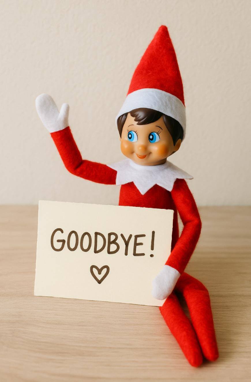 A red and white elf doll sits on a wooden surface, smiling and waving, while holding a sign that says GOODBYE! with a heart drawn underneath.
