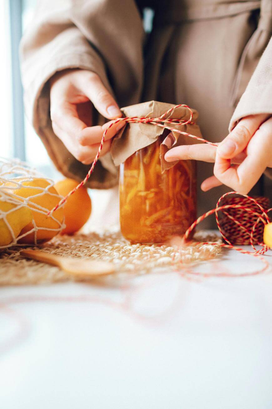 A person wraps twine around the top of a glass jar filled with preserved food, covering it with brown paper. Nearby are oranges in a net bag and a wooden spoon on a woven mat.