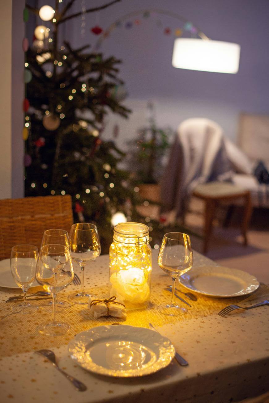 A warmly lit dining table set for four with wine glasses, plates, and cutlery. A mason jar filled with string lights is at the center. In the background, a decorated Christmas tree and cozy living room furniture are visible.