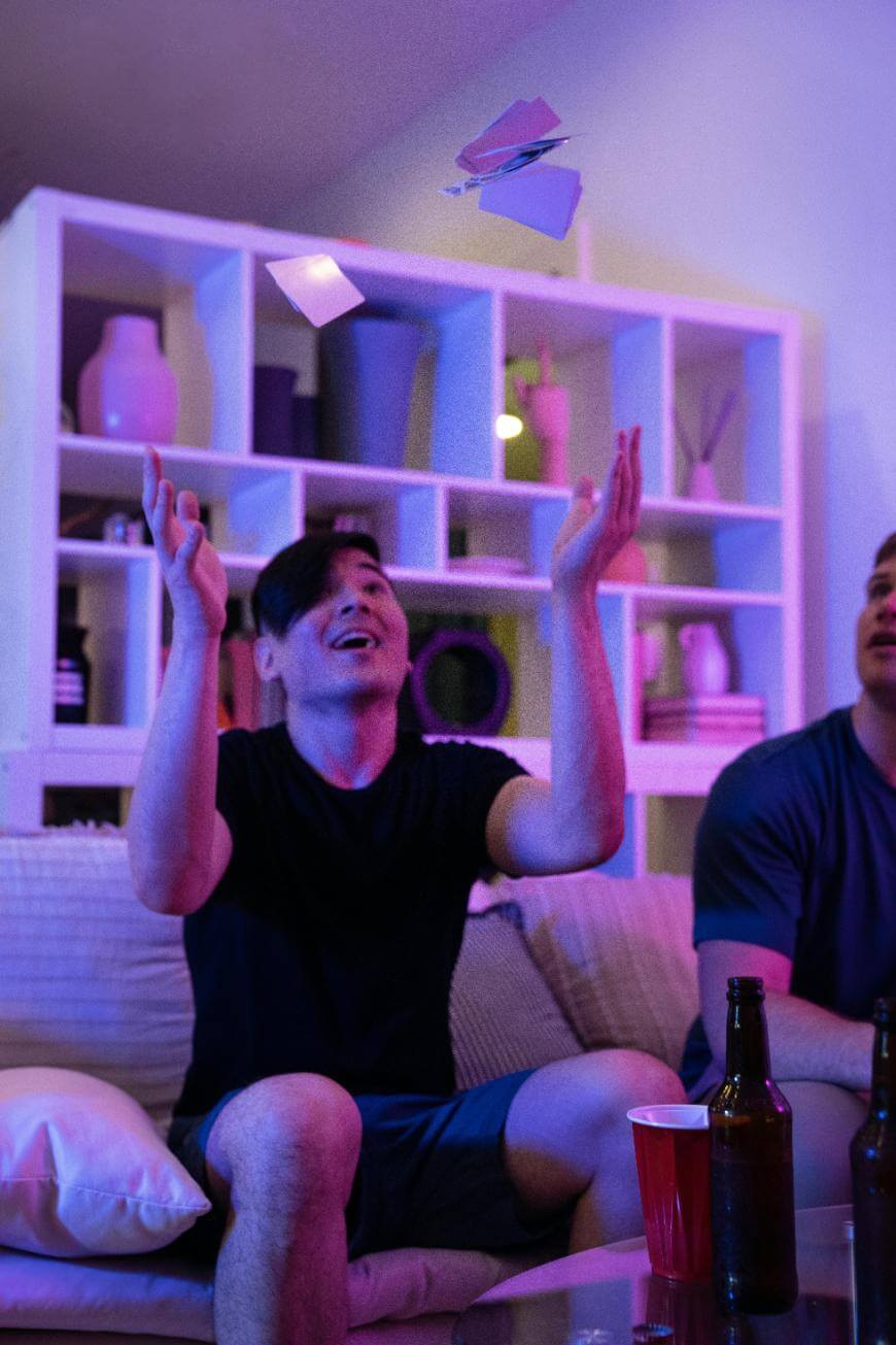 A man sitting on a couch tosses playing cards into the air, smiling, with colorful lighting, empty bottles, and a red cup on a table in front of him. Shelves with decor are visible in the background.