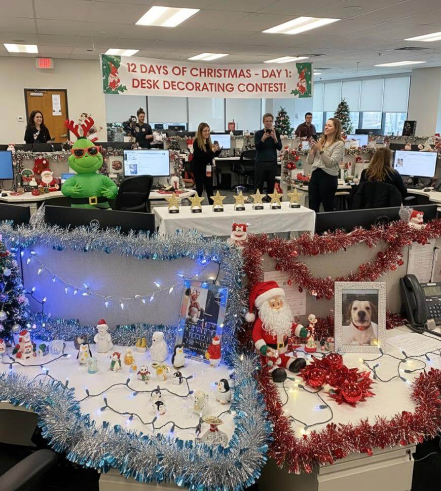 Office decked out for a Christmas desk decorating contest, with tinsel, lights, ornaments, Santa figures, and snowmen on desks. Staff stand and smile among festive decorations; a contest banner hangs above.