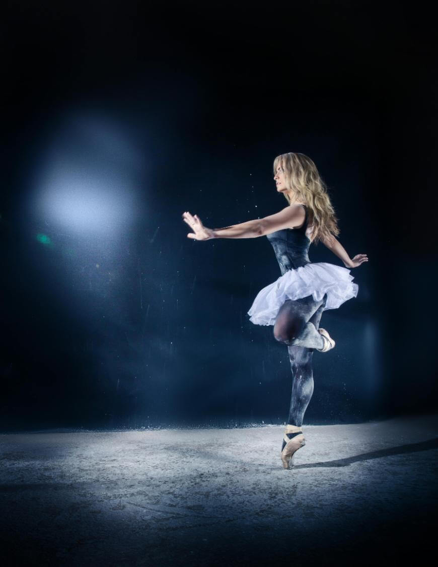 A ballet dancer in a black leotard and white tutu balances gracefully on one pointe, with one arm extended forward, against a dark, softly lit background.