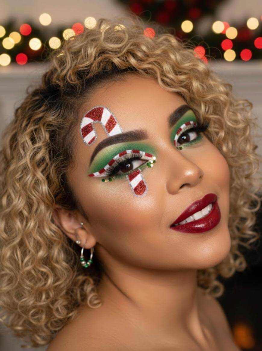 A woman with curly blonde hair wears bold holiday makeup with red lipstick, green eyeshadow, and candy cane designs above her eyes. She has festive earrings and blurred holiday lights in the background.