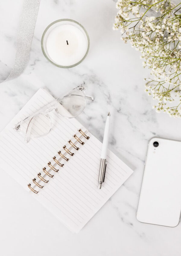Flat lay of a marble desk with an open lined notebook, a pen, eyeglasses, a white smartphone, a lit candle, a silver ribbon, and a bouquet of small white flowers.