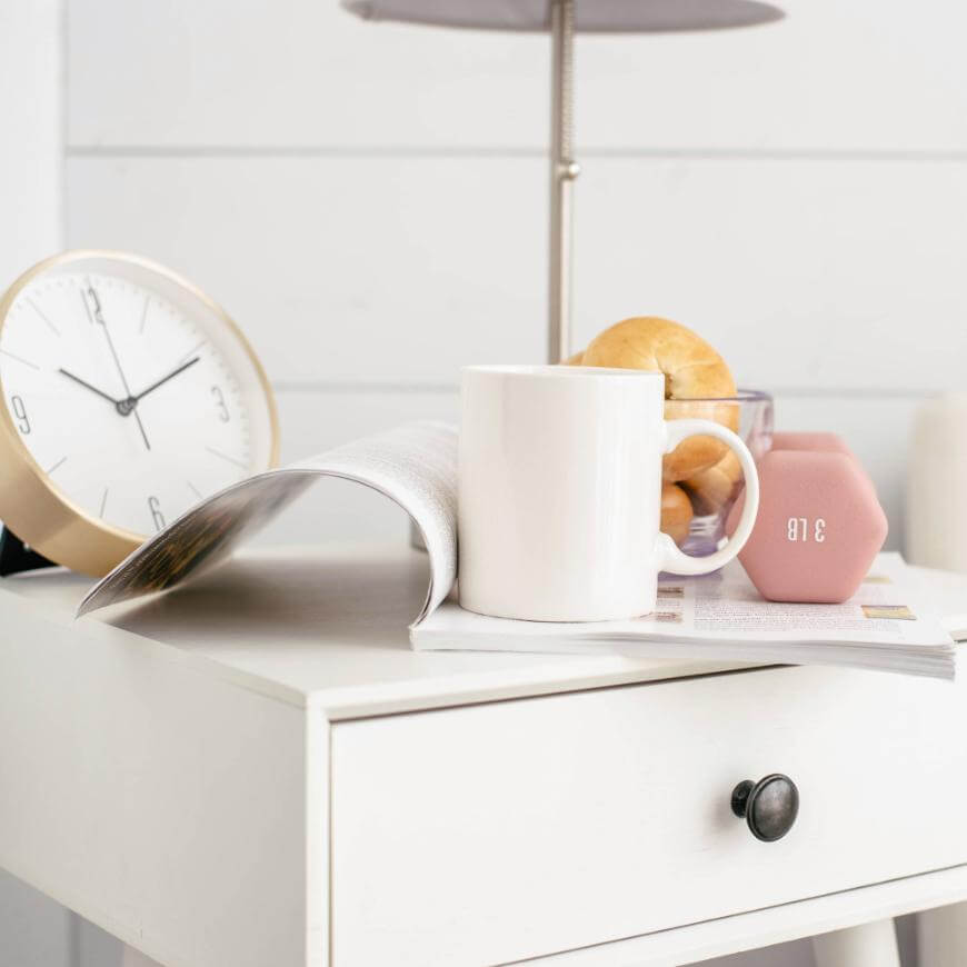 A white mug, an open magazine, a pink 3-pound dumbbell, two bread rolls, and a gold-rimmed clock sit on a white nightstand with a black knob, beneath a lamp.