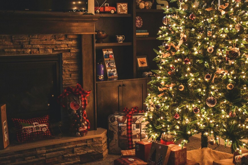 A decorated Christmas tree with glowing lights stands beside a fireplace and shelves, with wrapped presents and festive decorations arranged on the floor and mantle.