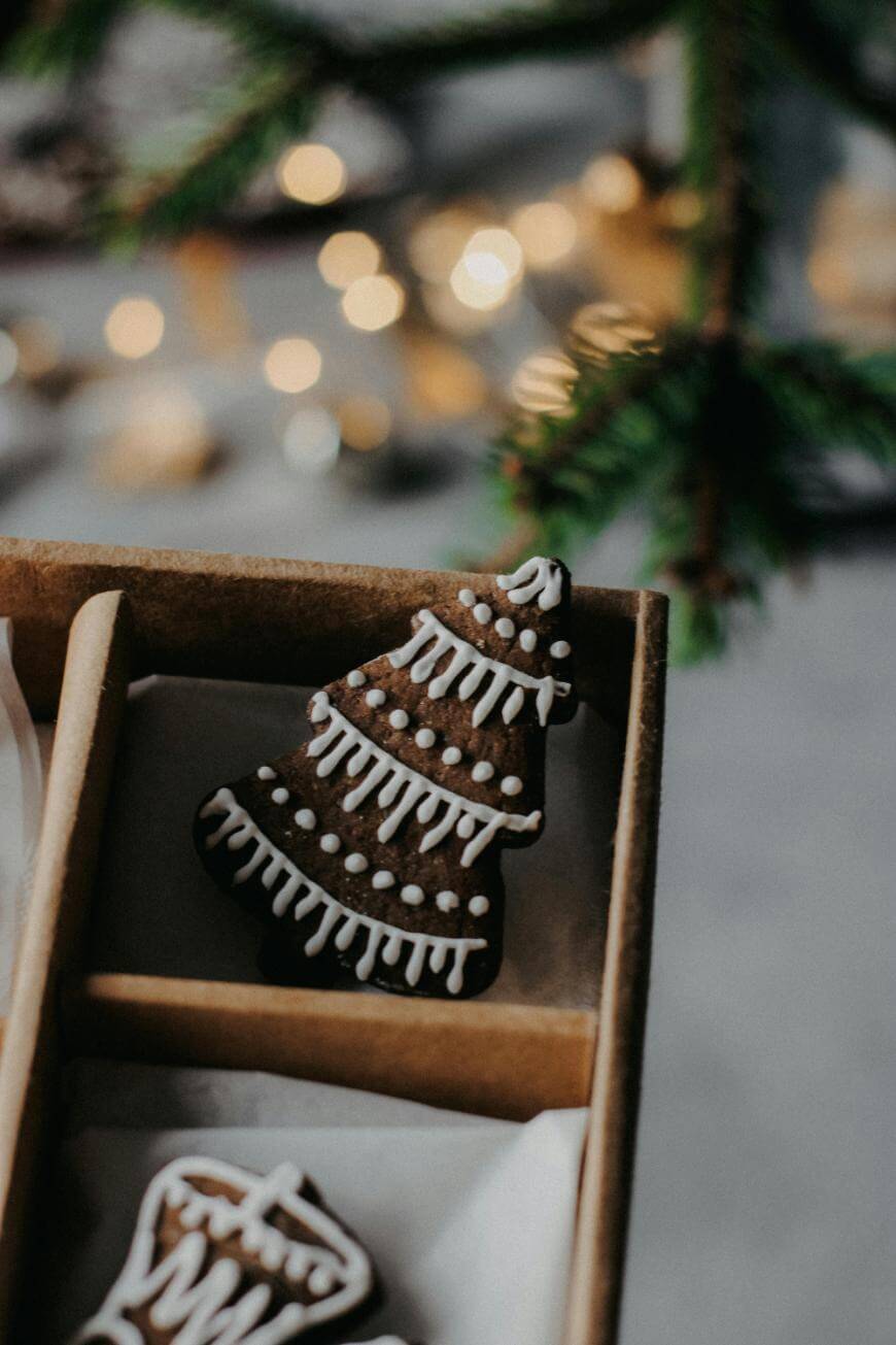 A gingerbread cookie shaped like a Christmas tree, decorated with white icing, sits in a brown box. Out-of-focus lights and a pine branch are visible in the background.