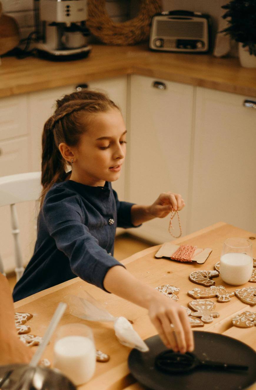 A young girl decorates gingerbread cookies at a kitchen table, surrounded by cookies, icing, a glass of milk, and sprinkles, creating a festive and cozy atmosphere.