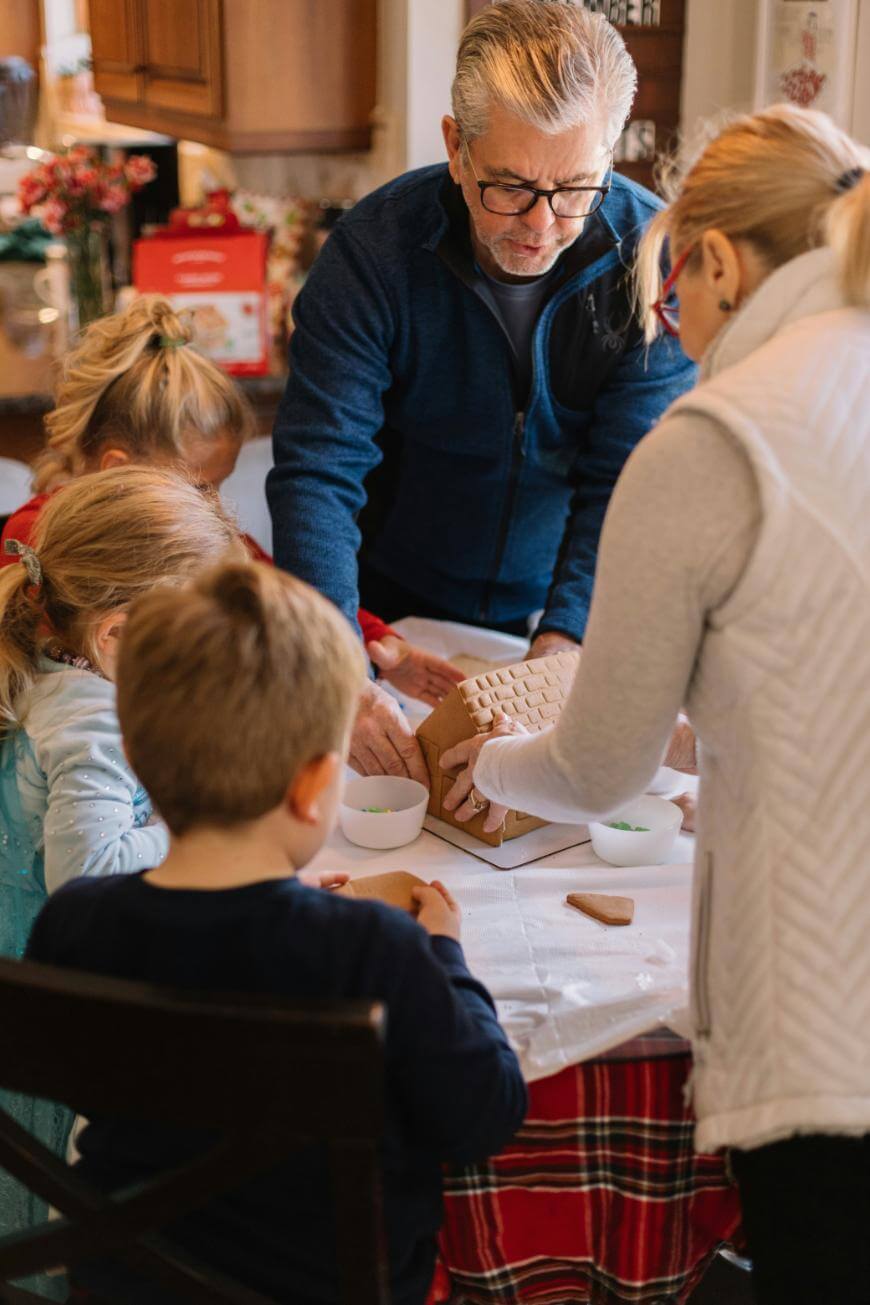A group of children and two adults gather around a table, decorating a gingerbread house together. The table is covered with a red plaid cloth and bowls of candy and icing are visible. The scene is warm and festive.