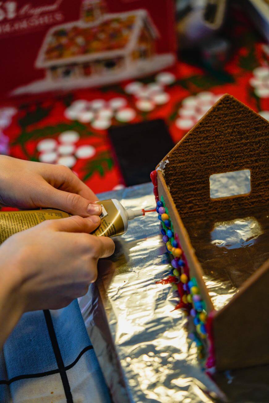 A person decorates a gingerbread house with colorful candies and frosting, using a tube of icing on a foil-covered surface. A festive red tablecloth and a gingerbread house kit box are in the background.