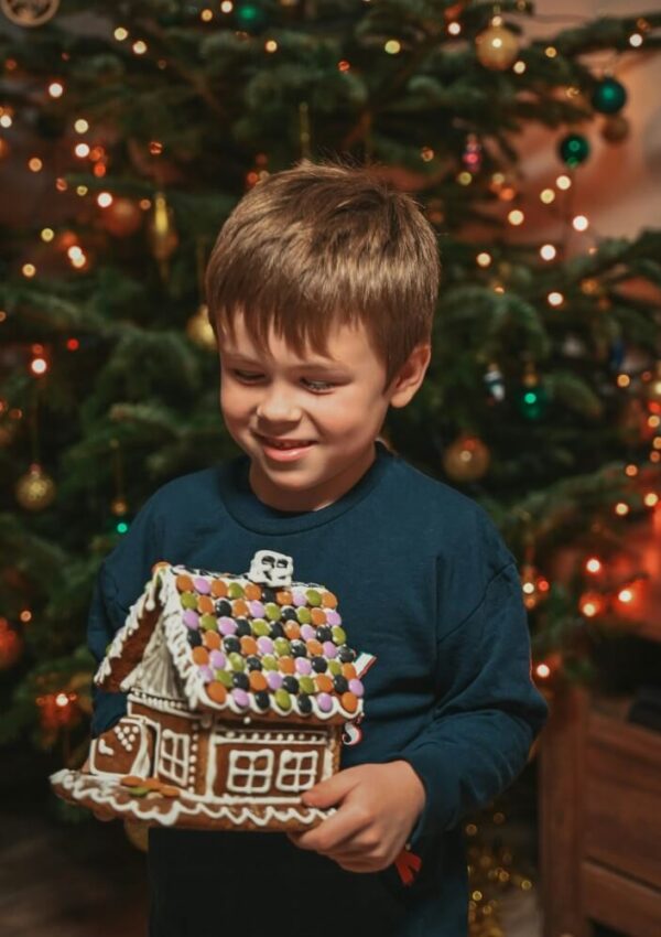 A young boy smiles while holding a decorated gingerbread house in front of a lit Christmas tree adorned with ornaments.