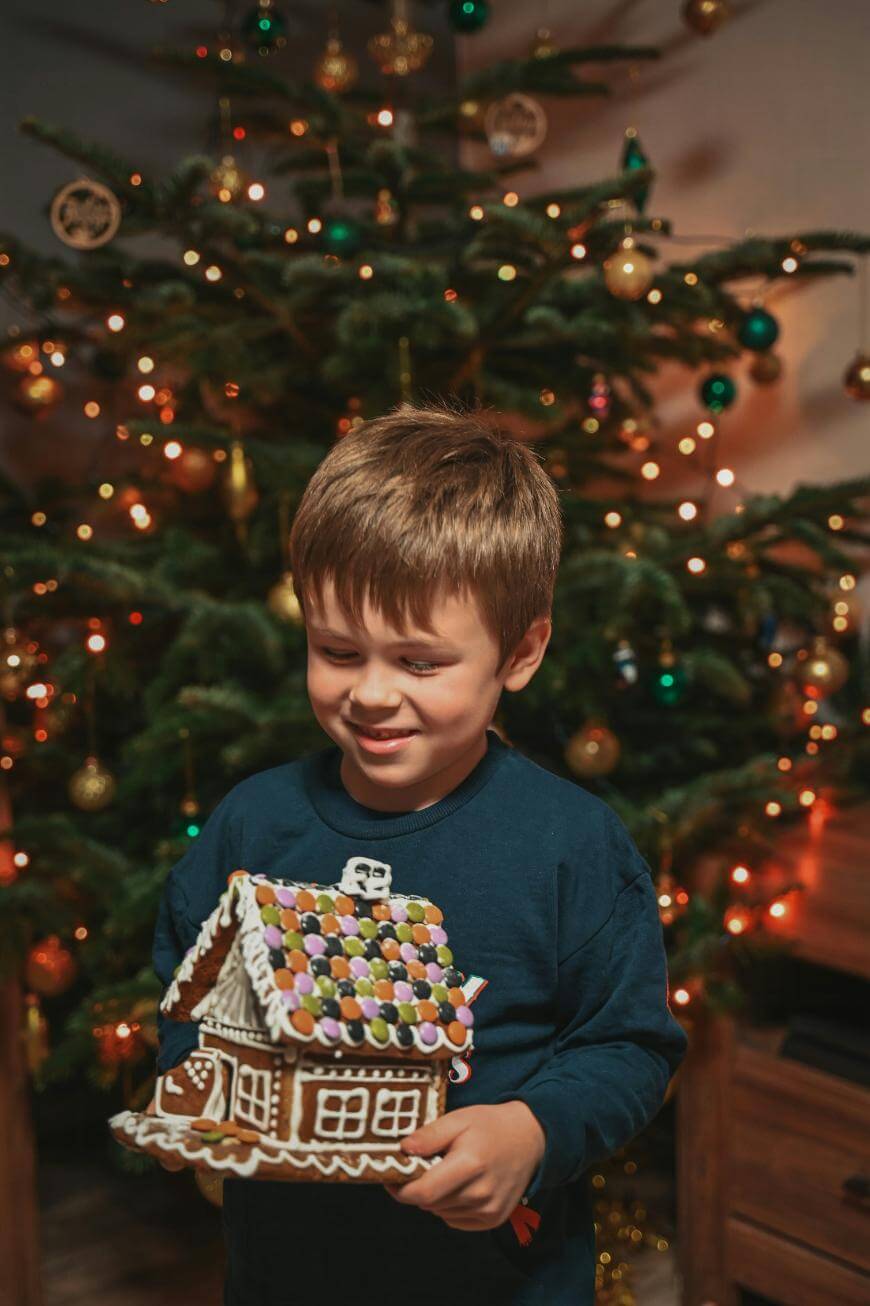A smiling young boy stands in front of a decorated Christmas tree, holding a gingerbread house covered with colorful candies.