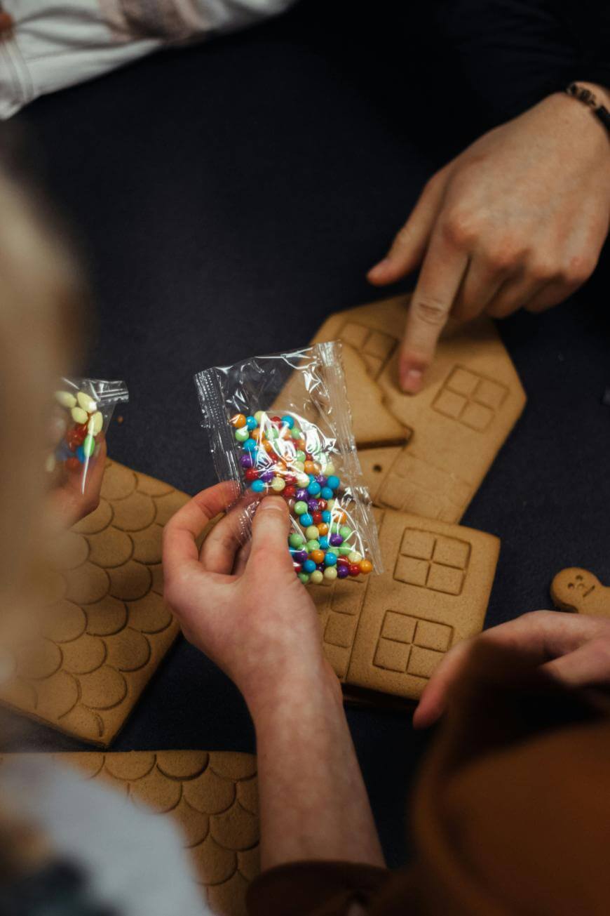 A person holds a packet of colorful candy while others point at unassembled gingerbread house pieces on a dark table.