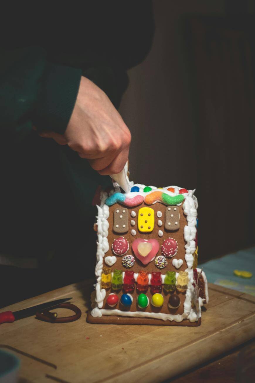 A person decorates a colorful gingerbread house with icing, adding details to the roof while the house stands on a wooden board. Various candies and sweets cover the house.