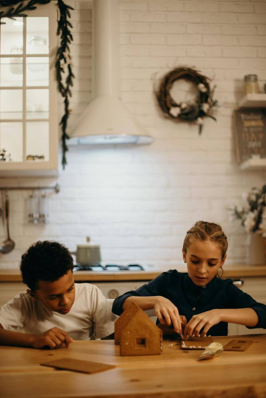 Two children sit at a kitchen table assembling a gingerbread house. The kitchen has white brick walls, hanging wreaths, and shelves with jars, creating a warm, festive atmosphere.