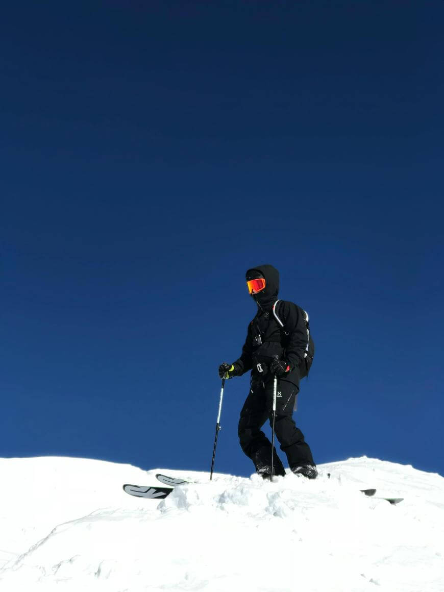 A skier dressed in black winter gear and orange goggles stands on a snowy slope against a clear blue sky, holding ski poles and looking into the distance.