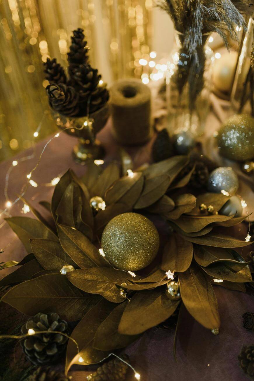 A festive table centerpiece with gold leaves, pinecones, glittery gold ornaments, small string lights, and decorative jars, creating a warm and elegant holiday atmosphere.