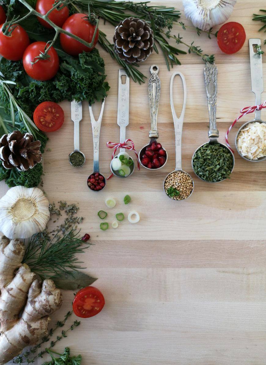 Measuring spoons with spices, herbs, pomegranate seeds, and green onions are arranged on a wooden surface, surrounded by fresh tomatoes, garlic, ginger, kale, pinecones, and sprigs of rosemary and thyme.
