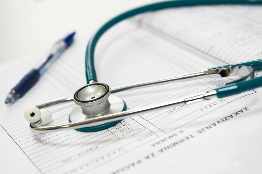 A teal stethoscope rests on top of a medical document with handwritten notes, alongside a blue pen.