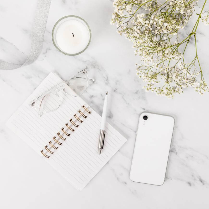 A flat lay on a white marble surface showing an open lined notebook with a pen, a pair of glasses, a white smartphone, a lit candle, and sprigs of white flowers.