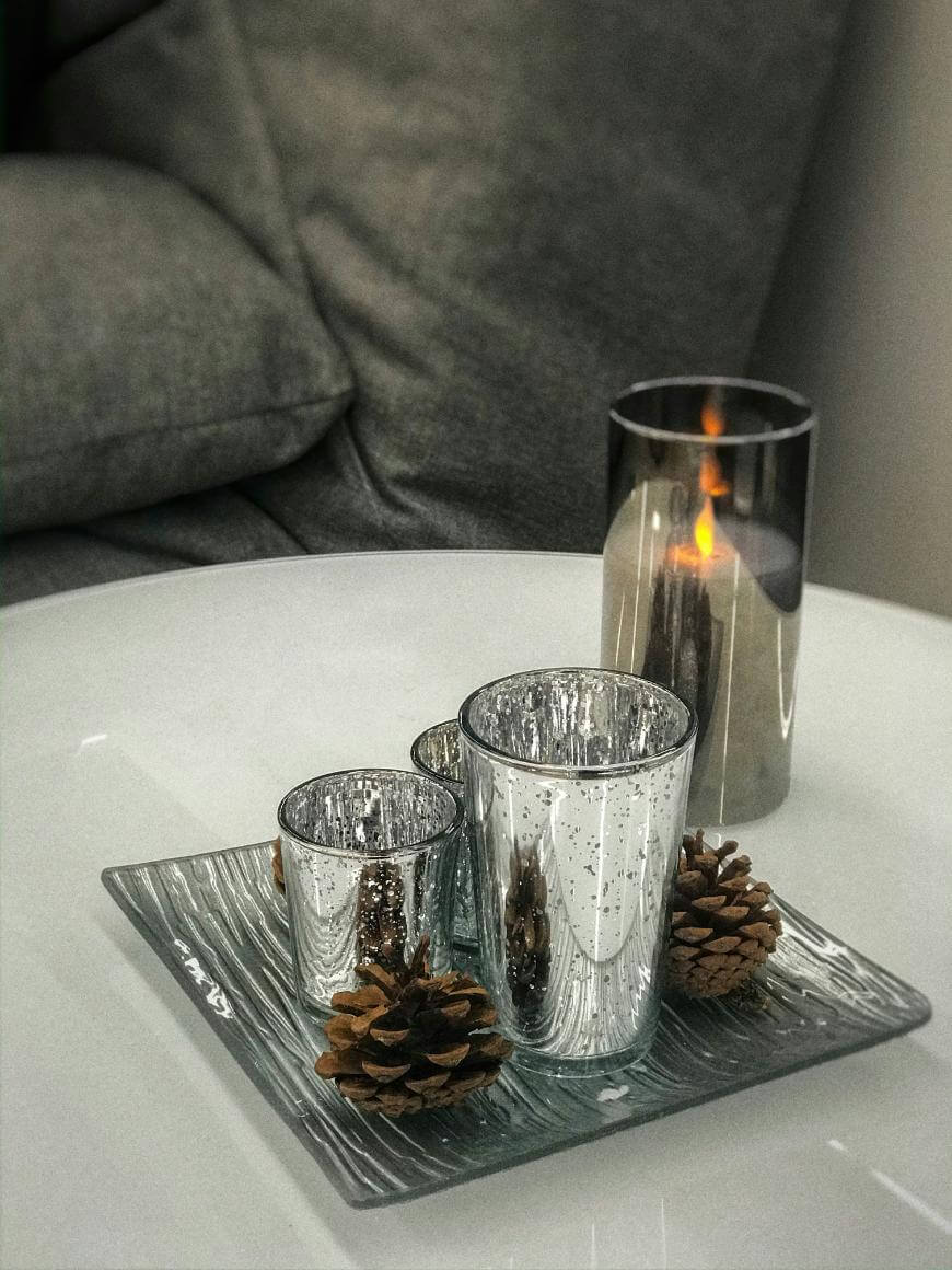 A white table with three silver votive candle holders and two pinecones on a glass tray, next to a lit candle in a dark glass holder. A gray sofa with cushions is in the background.