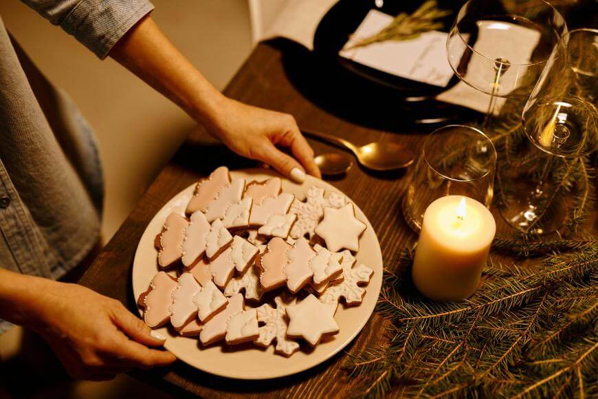 A person places a plate of iced cookies shaped like stars and Christmas trees on a wooden table set with a lit candle, pine branches, and glassware.