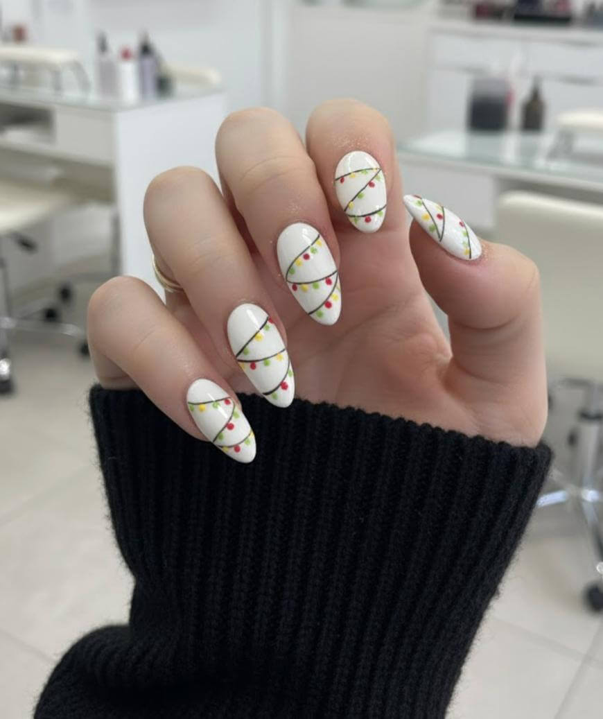 A hand with long, almond-shaped nails painted white, decorated with lines and small dots in red, green, and yellow, resembling festive holiday lights. The person is wearing a black sweater.