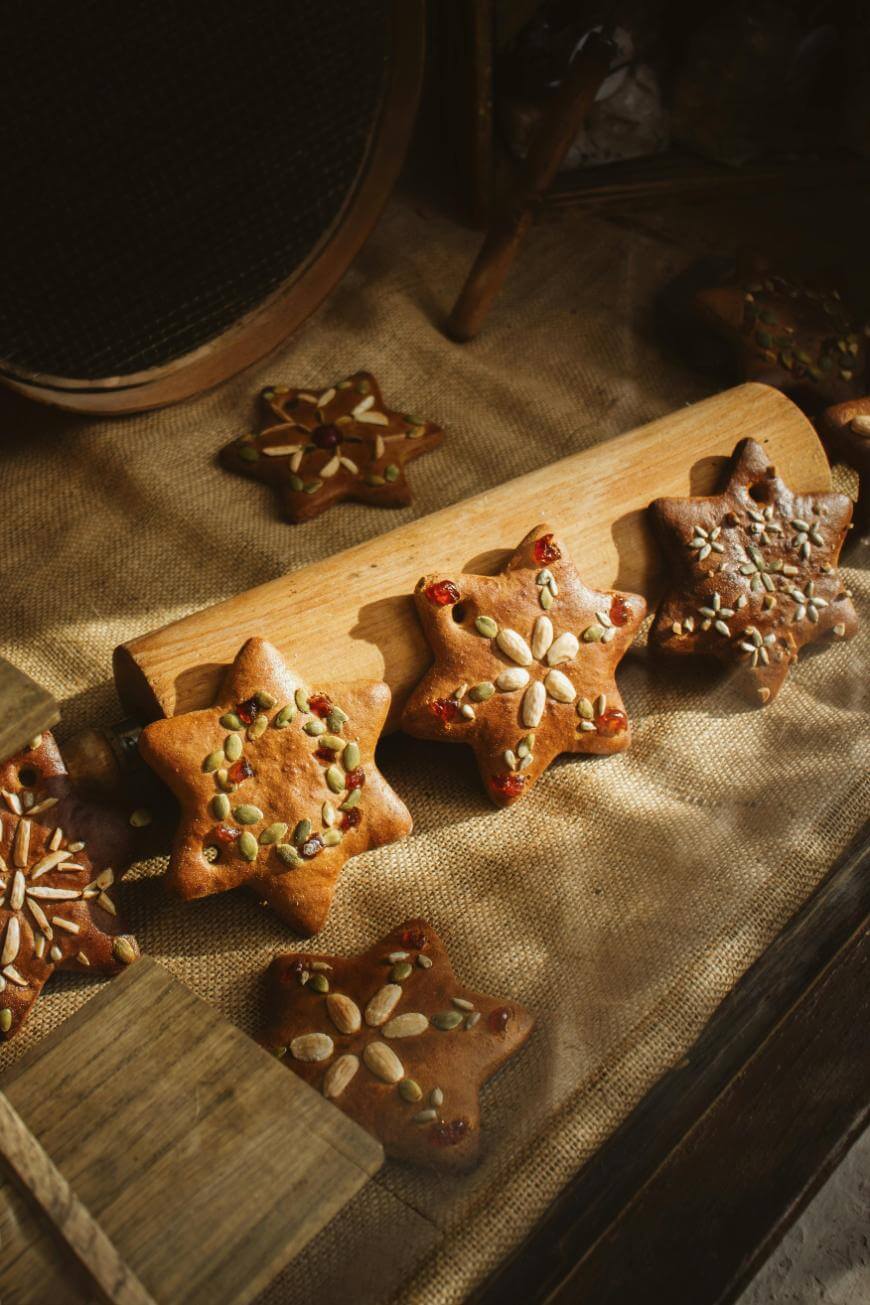 Star-shaped cookies decorated with various seeds and nuts are arranged on a wooden board and burlap surface, creating a rustic, homemade appearance in warm lighting.
