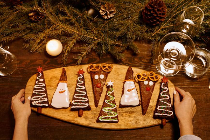 A person holds a wooden board with brownies decorated as Christmas trees and Santa faces. The table features pine branches, pinecones, a lit candle, empty glasses, and festive decor.