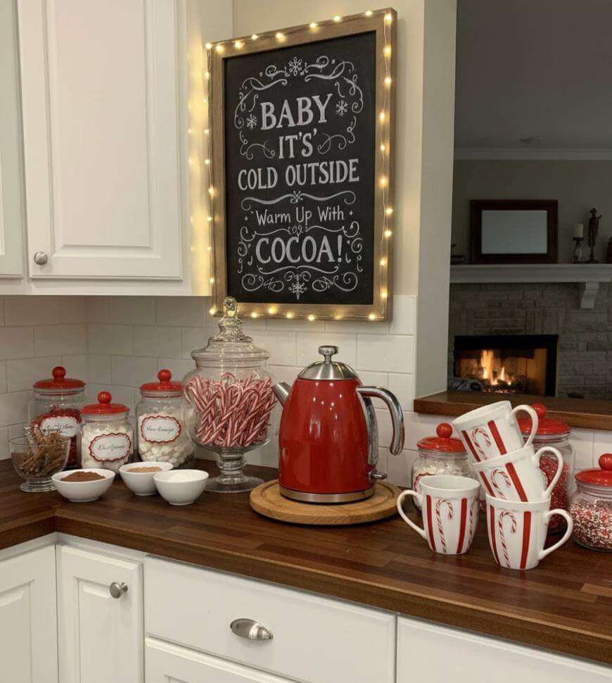 A cozy kitchen cocoa station with a red kettle, mugs with candy cane stripes, jars of cocoa mix, marshmallows, and candy canes on a wooden counter. A chalkboard sign reads, Baby its cold outside, warm up with cocoa!.